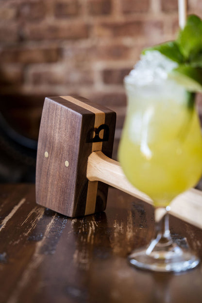 Diamond W Ranch black walnut ice mallet on a dark bar beside a yellow cocktail with mint, brick wall behind